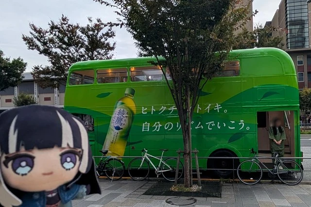 儒烏風亭らでんのぬいぐるみと、綾鷹『ヒトクチヒトイキ旅』バスの側面を撮影。綾鷹の写真とブランドメッセージが鮮やかに映る緑の車体。