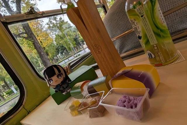 テーブルの上に並ぶ3種の和菓子と綾鷹、そして儒烏風亭らでんのぬいぐるみ。奥の窓からは秋のいちょう並木が見え、穏やかな『ヒトクチヒトイキ旅』のひと幕を切り取った一枚。