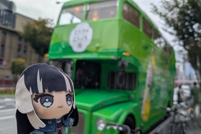 儒烏風亭らでんのぬいぐるみと、綾鷹コラボ『ヒトクチヒトイキ旅』バスの正面を背景に撮影した写真。緑色の車体が印象的。
